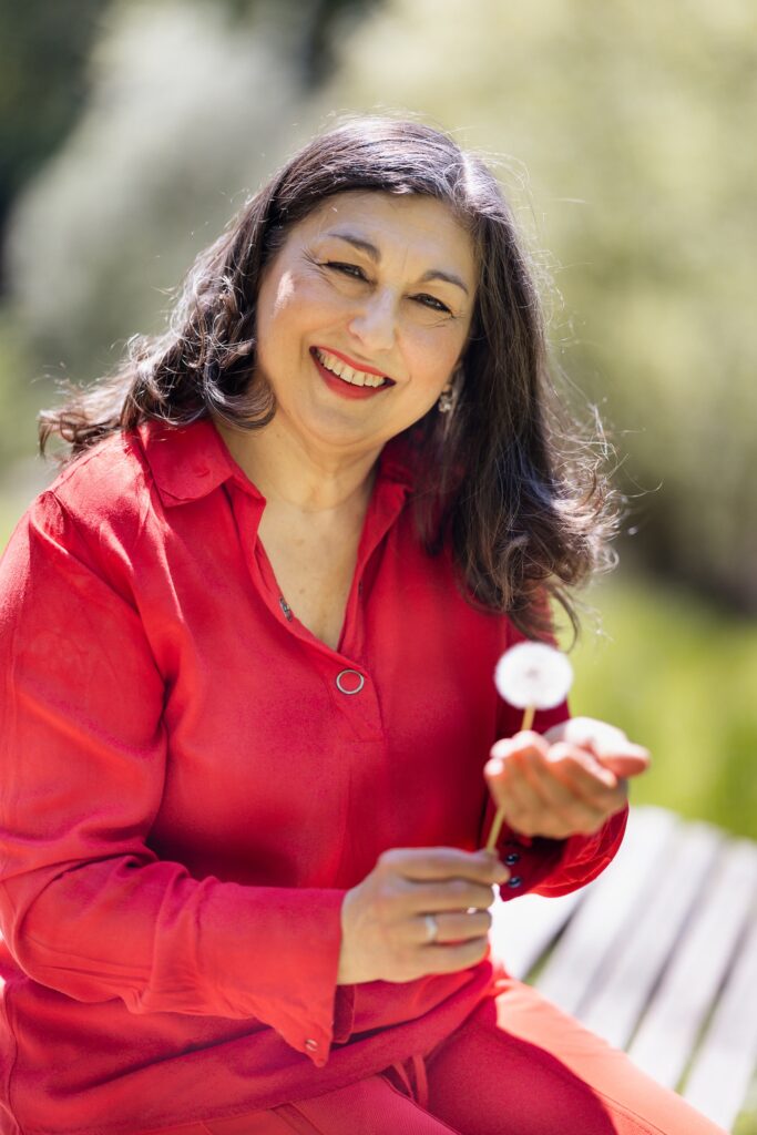 Carmen Uth im roten Oberteil laechelnd mit Pusteblume in der Hand – Sinnbild fuer Klarheit, emotionale Staerke und achtsames Coaching.